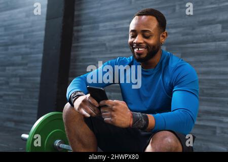 Junger Sportler, der Smartphone benutzt, sitzt auf der Langhantel im Fitnessstudio Stockfoto