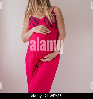 Eine anspruchsvolle Schwangere in einem rosa Kleid steht auf weißem Hintergrund mit den Händen auf dem Bauch, dem Konzept der Weiblichkeit Stockfoto