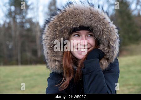Lächelnde junge Frau mit warmer Pelz-Kapuzenjacke, die in die Natur blickt Stockfoto