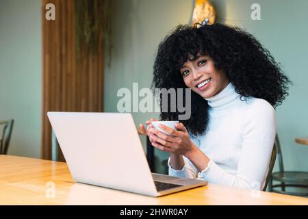 Lächelnde, lockige, schwarzhaarige Frau mit Kaffeetasse, die mit einem Laptop im Café sitzt Stockfoto