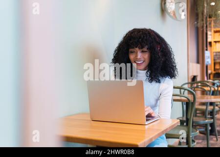 Lächelnde junge Frau mit lockigen Haaren, die am Tisch im Café einen Laptop benutzt Stockfoto