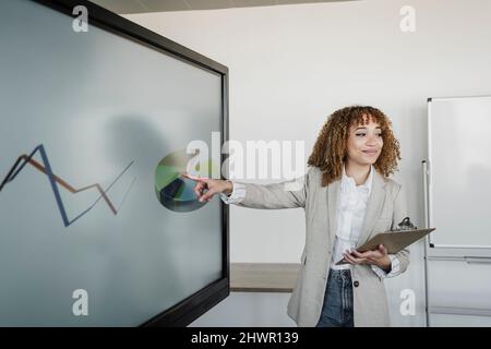 Lächelnde junge Frau mit braunem lockigen Haar, die auf Pie hart im Konferenzraum zeigt Stockfoto