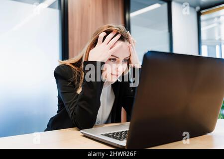 Frustrierte Geschäftsfrau mit Kopf in den Händen und Blick auf den Laptop im Büro Stockfoto