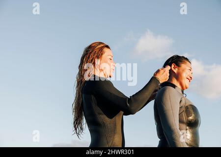 Lächelnde Frau, die der Surferin beim Anziehen von Neoprenanzüge am Strand assistierte Stockfoto