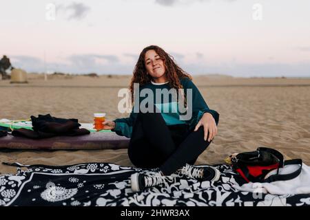 Junge Frau mit Bierbecher auf Sand am Strand sitzen Stockfoto