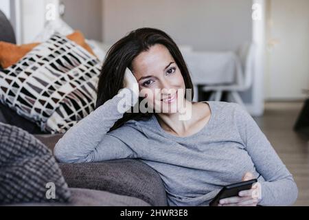 Lächelnde Frau mit Smartphone, das zu Hause auf dem Sofa gelehnt ist Stockfoto
