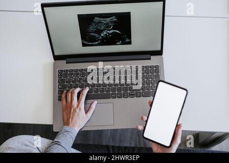 Frau, die zu Hause Smartphone mit Ultraschall auf dem Laptop hält Stockfoto