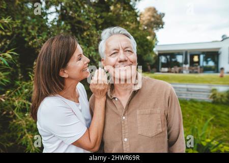 Lächelnde Frau flüstert im Hinterhof in das Ohr des älteren Mannes Stockfoto