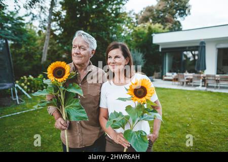 Lächelnder älterer Mann mit Frau, die Sonnenblumen im Hinterhof hält Stockfoto