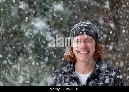 Lächelnder junger Mann in Strickmütze, der Schneefall genießt Stockfoto
