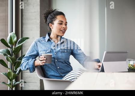 Junge Geschäftsfrau mit Kaffeetasse, die im Büro am Tablet-Computer arbeitet Stockfoto