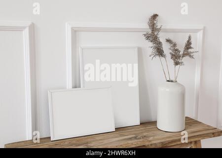 Satz von weißen Bilderrahmen-Mockups. Vase mit trockenem Schilf, Gras auf alter Holzbank. Wandverzierungen im Hintergrund, Zierleisten. Elegante Inneneinrichtung Stockfoto