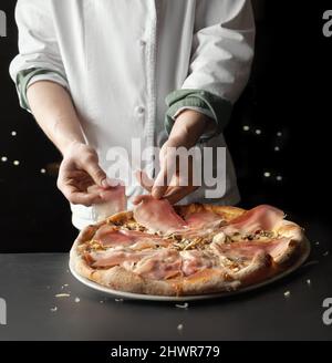 Der Küchenchef bereitet Pizza mit Prosciutto crudo und Parmesan auf schwarzem Hintergrund zu. Stockfoto