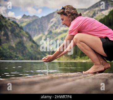 Frau, die Wasser in geschöpften Händen hält und am Seeufer hockt Stockfoto