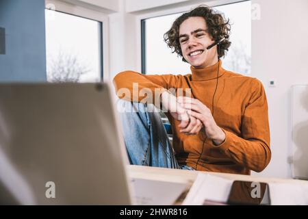 Ein glücklicher junger Student, der Kopfhörer trägt und zu Hause über einen Laptop lernt Stockfoto
