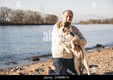 Lächelnder älterer Mann umarmte den Hund am Strand an einem sonnigen Tag Stockfoto