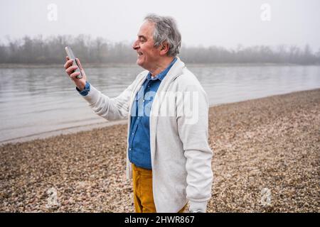 Lächelnder älterer Mann, der am Strand einen Videoanruf über das Mobiltelefon führt Stockfoto