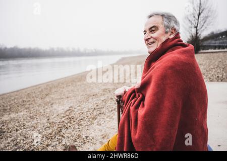 Glücklicher Mann, der in eine Decke gehüllt am Strand sitzt Stockfoto