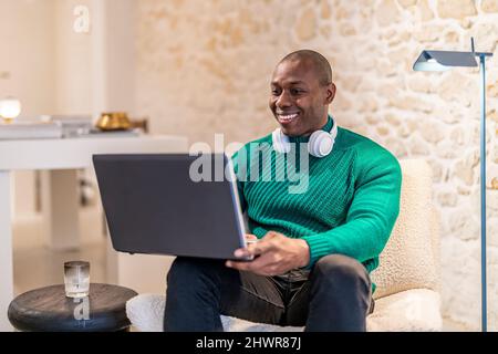 Glücklicher Mann mit kabellosen Kopfhörern, der mit einem Laptop zu Hause arbeitet Stockfoto