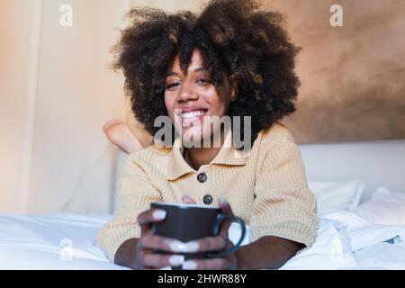 Lächelnde Frau mit Kaffeetasse, die im Schlafzimmer auf dem Bett lag Stockfoto
