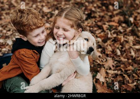 Happy girl umarmt golden Retriever Welpen von Bruder auf Herbstblätter im Wald Stockfoto