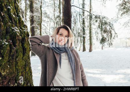 Lächelnde Frau, die im Winterwald mit Händen hinter dem Kopf an einem Baum steht Stockfoto