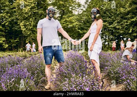 Junges Paar, das in einem Lavendelfeld steht und Gasmasken trägt und die Hände hält. Menschen mit Allergien und blühenden Lavendelblüten. Konzept der saisonalen Allergie Stockfoto