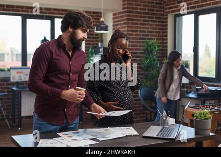 Die werdende Mutter telefoniert im Büro der Finanzagentur, während die Kollegin der Abteilung wichtige Unterlagen liest. Mitarbeiter des Unternehmens prüft Ideen zur Projektmarketingstrategie. Stockfoto