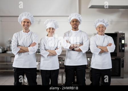 Porträt einer jungen, multirassischen Gruppe von Köchen, die in Uniformen in der Restaurantküche zusammenstehen. Selbstbewusste Köche, die bereit sind, im Restaurant zu kochen. Team aus europäischen, asiatischen und lateinamerikanischen Fachleuten Stockfoto