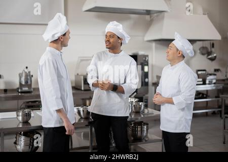 Drei Köche aus verschiedenen Rassen unterhalten sich während einer Pause in der Restaurantküche, während sie zusammen stehen. Asiatische, lateinamerikanische und europäische Köche tragen weiße Uniformen bei der Arbeit Stockfoto
