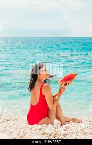 Junge hübsche Frau in rotem Badeanzug am Strand mit Wassermelone-Kopierfläche Stockfoto
