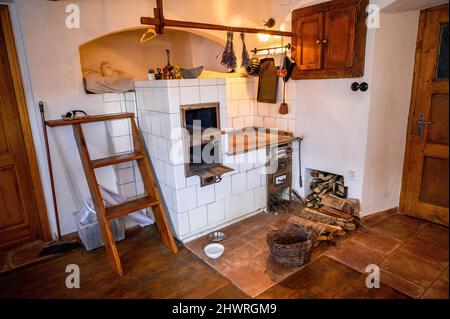 Alte ländliche Küche mit weißem Keramik-Kamin Herd und Backofen, Holz, Korb und verschiedene Utesil im Dorfhaus, Tschechische republik. Stockfoto