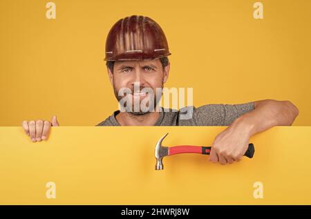 Glücklicher reifer Mann Arbeiter im Helm hinter gelbem Papier für Kopierraum halten Hammer, glücklicher Arbeiter Tag Stockfoto
