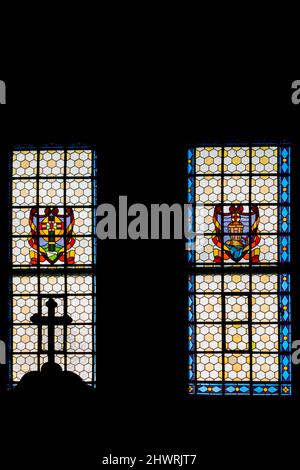 Das katholische Kreuz auf der Spitze des Beichtstuhls vor dem Hintergrund eines sonnendurchfluteten Buntglasfensters. Das Foto wurde in der Kirche aufgenommen. Stockfoto