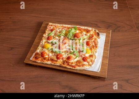 Viereckige Pizza mit zweifarbigen Kirschtomaten, Rucola und Mozzarella-Käse mit Orangano auf einem Holztisch Stockfoto