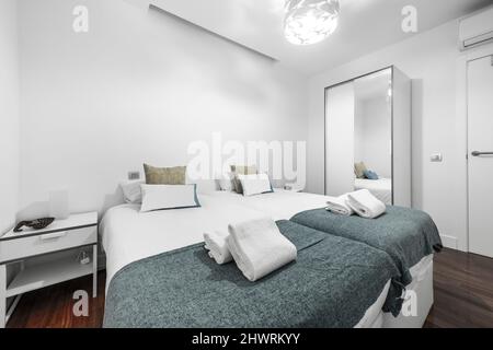Schlafzimmer mit Doppelbetten mit weißen Bettdecken, passenden Jalousien, kleinen blauen Decken und Nachttischen mit zwei Lampen, Kleiderschrank mit verspiegelter Tür in VA Stockfoto
