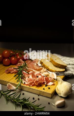 Bio-Salami, Schweinehals, trockener Schinken mit Kirschtomaten, Rosmarinzweig, Knoblauch und rustikalem Brot auf einem Holzschneidebrett. Dunkler Hintergrund. Stockfoto