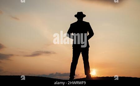 Silhouette eines Mannes im Hut, der am Sonnenaufgangshimmel steht, Einsamkeit Stockfoto