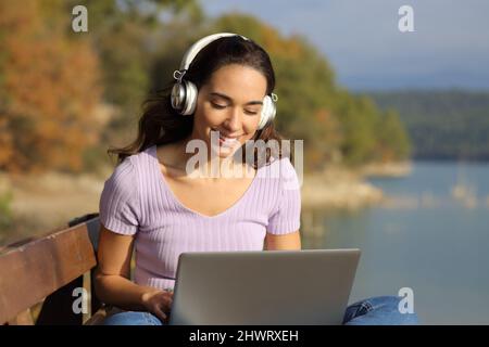 Glückliche Frau mit schnurlosem Headset, die den Laptop in der Natur überprüft Stockfoto