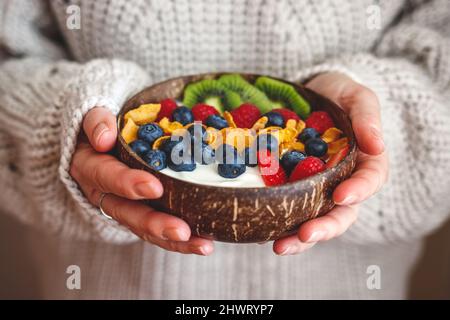 Gesundes Frühstück. Frau hält eine Kokosnussschale mit Joghurt, Cornflakes, Kiwi in Scheiben und Beerenobst. Diät-Nahrung für Wohlbefinden und gute Vitalität Stockfoto