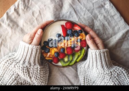 Gesundes Frühstück in Kokosnussschale auf dem Tisch mit Leinentischdecke. Joghurt mit Maisflocken und Heidelbeere, Erdbeere, Kiwi und Himbeere. Womans Hand Stockfoto