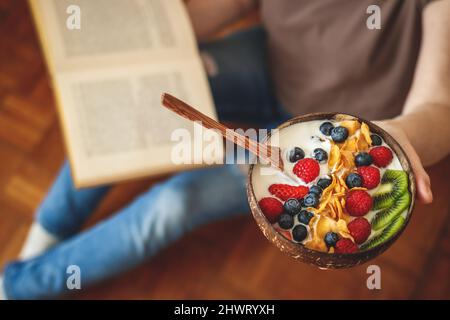 Joghurt, Obst und Cornflakes in Kokosnussschale. Frau liest Buch während des Frühstücks zu Hause. Gesunde Ernährung und Lebensstil Stockfoto