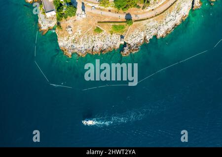 Die Yacht segelt in der Adria und hinterlässt einen weißen Seeweg in der Nähe des felsigen Strandes von Rovinj. Die Azure Bucht, die die kroatische Stadt bei hellem Sonnenlicht umgibt. Luftaufnahme Stockfoto