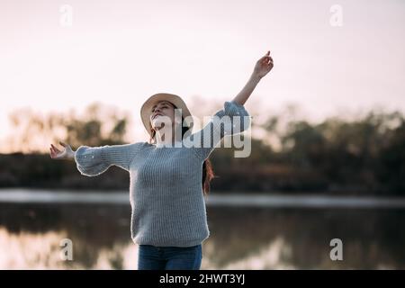 Die Brünette fühlt sich frei am Ufer eines Flusses, mit einem Hut, der ihre Arme hebt Stockfoto