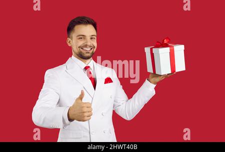 Glücklicher, gutaussehender Mann im Anzug, lächelnd, hält die Geschenkbox und zeigt die Daumen hoch Stockfoto