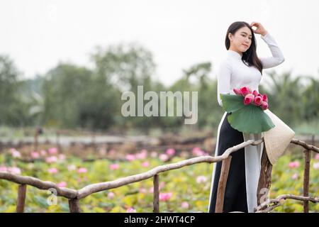 Porträt einer schönen vietnamesischen Frau mit traditionellem vietnam-Hut, der den rosa Lotus hält, der auf der Holzbrücke im großen Lotussee, vietnam, ai, läuft Stockfoto