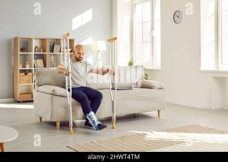 Der junge Mann mit Beinverletzung lehnt sich auf Krücken, während er zu Hause versucht, vom Sofa aufzustehen. Stockfoto