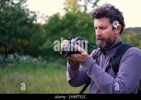 Ein männlicher Fotograf mit einer Kamille im Haar fotografiert etwas mit einer Fotosammlung in den Händen. Im Frühling Stockfoto