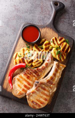 Schweinekoteletts mit gegrillten Kartoffeln, Sauce und Chilischoten aus der Nähe auf einem Holzbrett auf dem Tisch. Vertikale Draufsicht von oben Stockfoto
