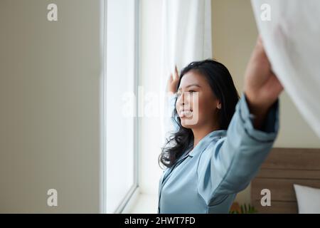 Glückliche junge Frau in seidenen Loungewear, die Vorhänge öffnet und durch das Fenster schaut und den neuen Morgen genießt Stockfoto